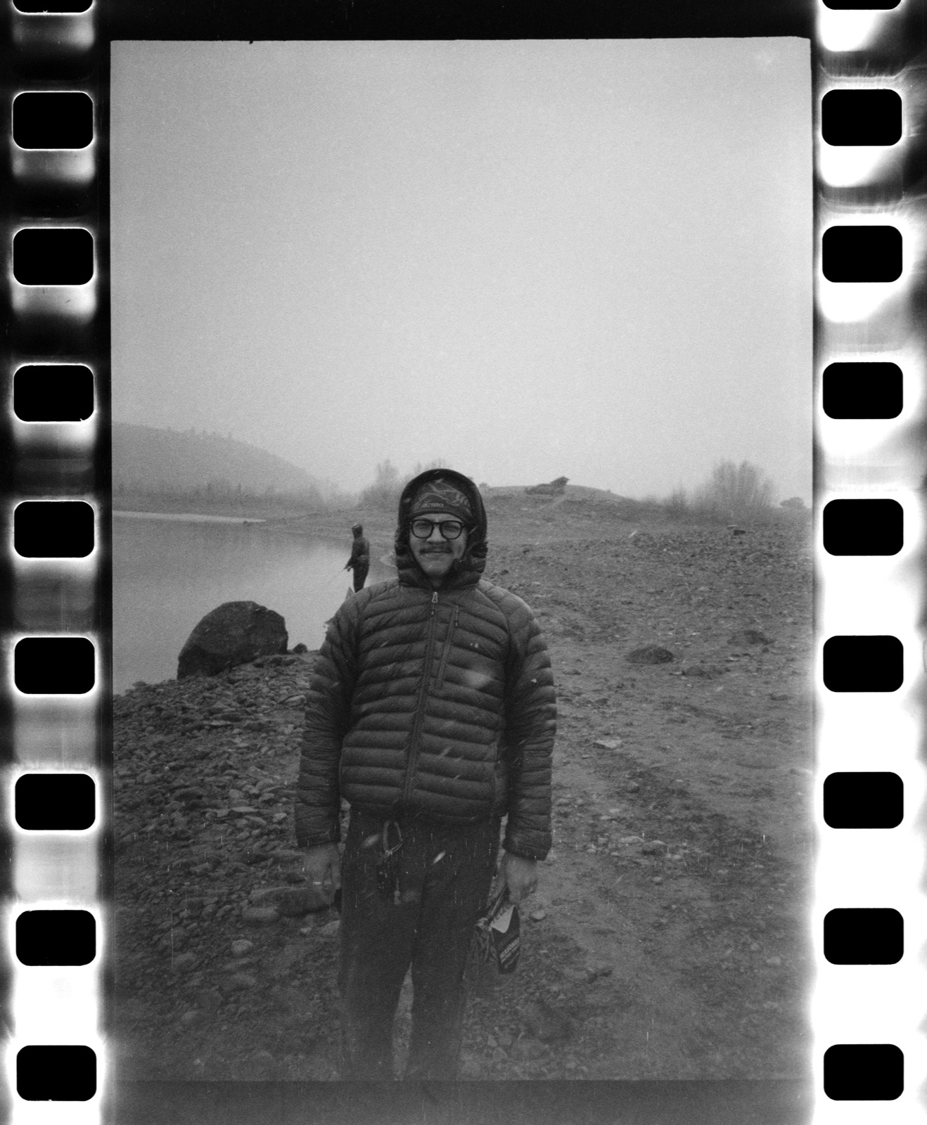 Judah in a hooded puffer jacket on a foggy, rocky riverbank, black and white film